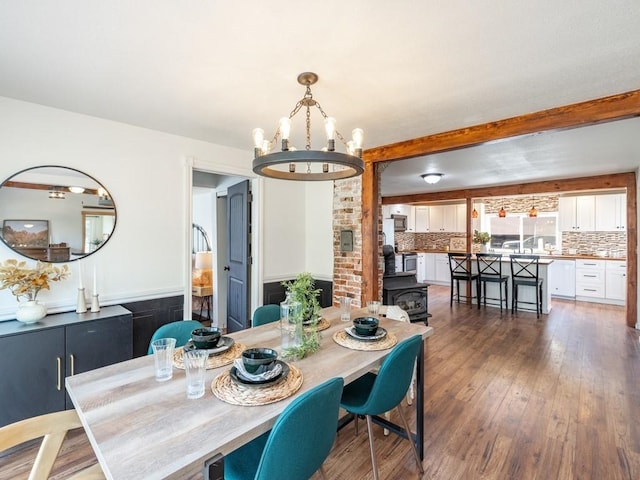 dining room featuring an inviting chandelier, a wood stove, beamed ceiling, wainscoting, and wood-type flooring