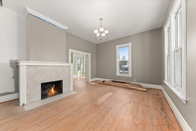unfurnished living room with baseboards, an inviting chandelier, a tiled fireplace, a wealth of natural light, and wood finished floors