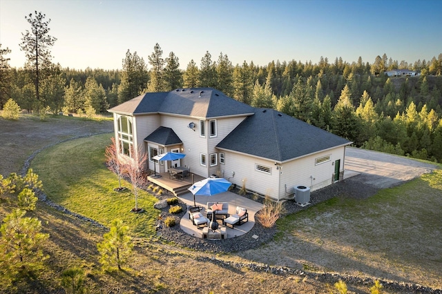 rear view of house featuring roof with shingles, a patio area, a forest view, a wooden deck, and a yard