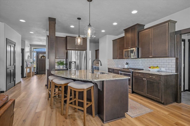 kitchen with a kitchen breakfast bar, dark brown cabinets, light wood-style floors, light stone countertops, and appliances with stainless steel finishes