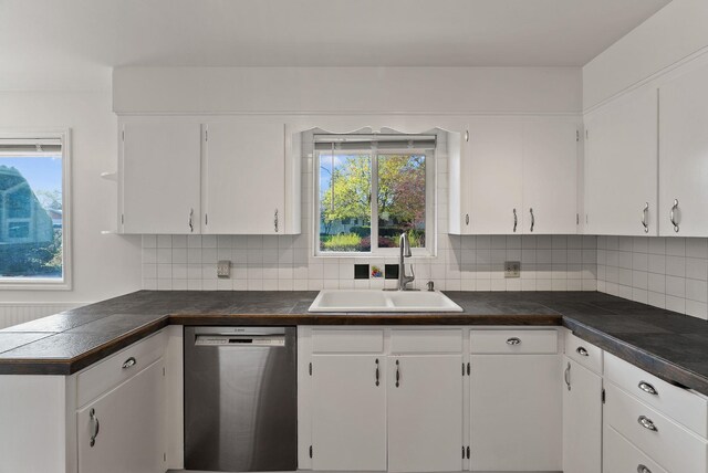 kitchen with dark countertops, stainless steel dishwasher, a sink, decorative backsplash, and white cabinetry