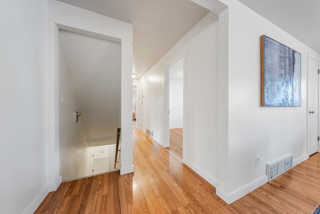 hall with light wood-style flooring, visible vents, baseboards, and an upstairs landing