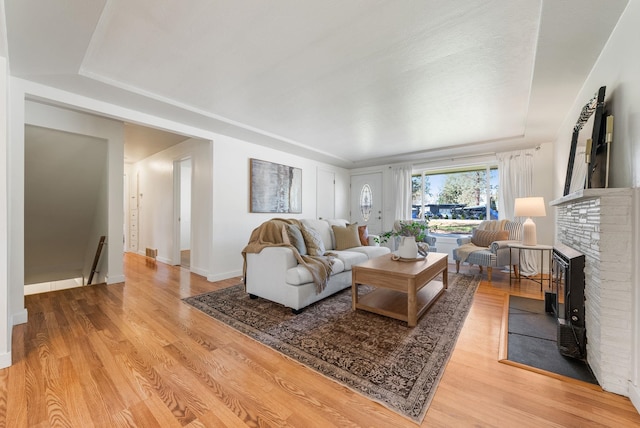 living area featuring baseboards, a brick fireplace, light wood-style floors, and a raised ceiling