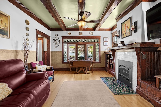 living room featuring a fireplace with flush hearth, beamed ceiling, ceiling fan, wood finished floors, and wainscoting