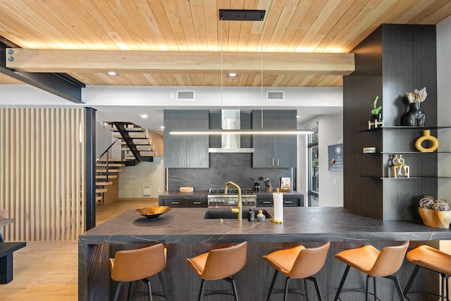 kitchen featuring wall chimney range hood, a kitchen breakfast bar, wood ceiling, dark countertops, and light wood finished floors