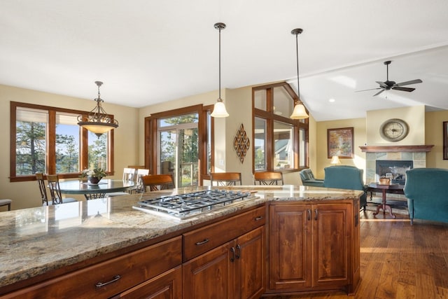 kitchen featuring a ceiling fan, a fireplace, stainless steel gas cooktop, light stone counters, and dark wood-style floors