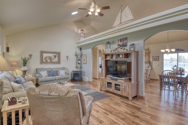 living room featuring a wood stove, light wood-style floors, high vaulted ceiling, arched walkways, and ceiling fan with notable chandelier
