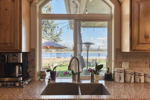 kitchen featuring decorative backsplash, a water view, brown cabinets, light stone counters, and a sink