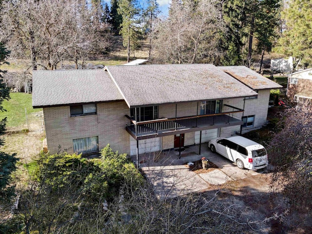 rear view of house featuring a balcony, brick siding, a shingled roof, an attached garage, and driveway