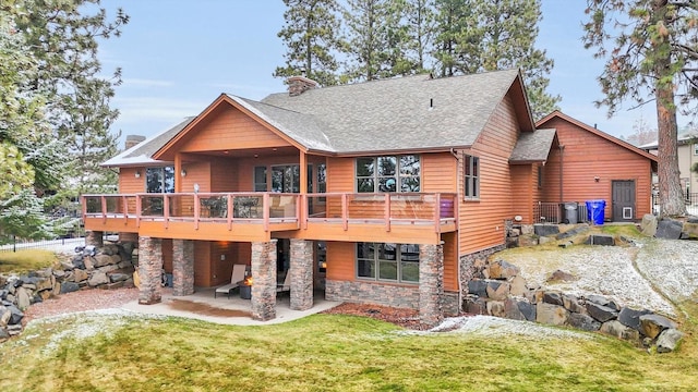 rear view of house featuring roof with shingles, stone siding, a wooden deck, a patio area, and a lawn