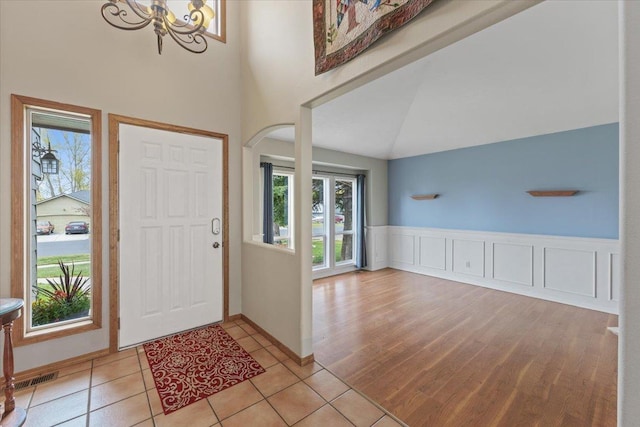 entryway with arched walkways, wainscoting, high vaulted ceiling, a chandelier, and light wood-style floors