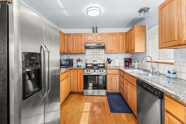kitchen featuring light stone countertops, a sink, under cabinet range hood, stainless steel appliances, and light wood-style flooring