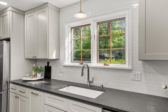 kitchen featuring dark countertops, stainless steel refrigerator, a sink, backsplash, and pendant lighting