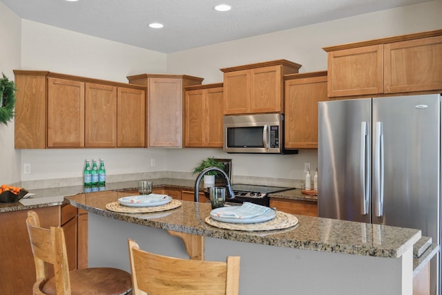 kitchen featuring a kitchen island with sink, stone countertops, brown cabinetry, stainless steel appliances, and a kitchen bar