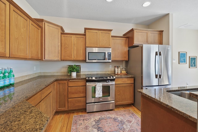 kitchen featuring light wood finished floors, brown cabinetry, dark stone counters, stainless steel appliances, and recessed lighting