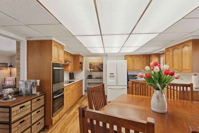 kitchen featuring white refrigerator with ice dispenser, double oven, black electric cooktop, a drop ceiling, and light wood-type flooring