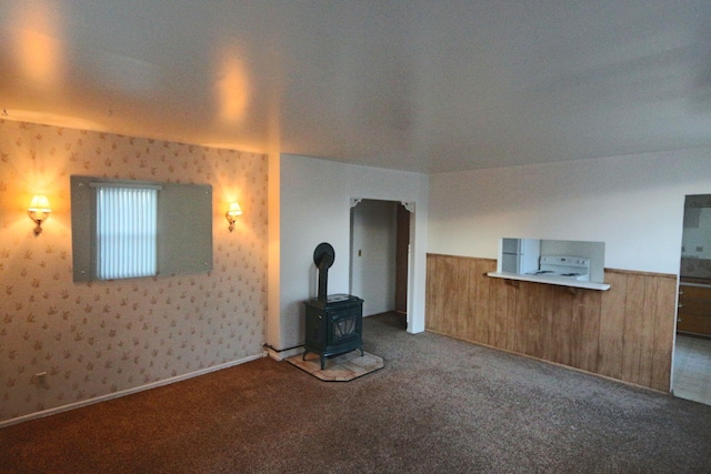 unfurnished living room featuring carpet floors, wallpapered walls, a wood stove, and a wainscoted wall
