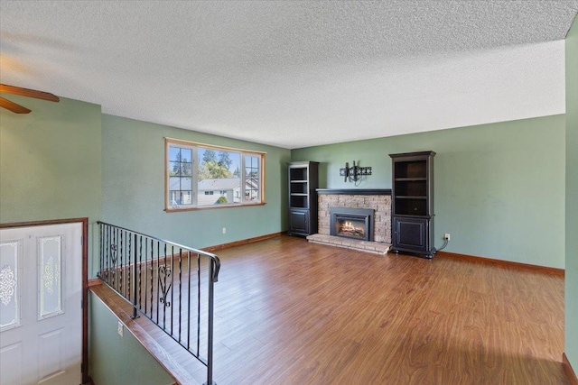unfurnished living room with a textured ceiling, a stone fireplace, a ceiling fan, baseboards, and wood finished floors