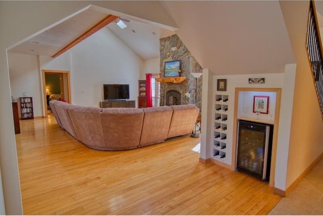 living area featuring wine cooler, a stone fireplace, light wood finished floors, high vaulted ceiling, and beam ceiling