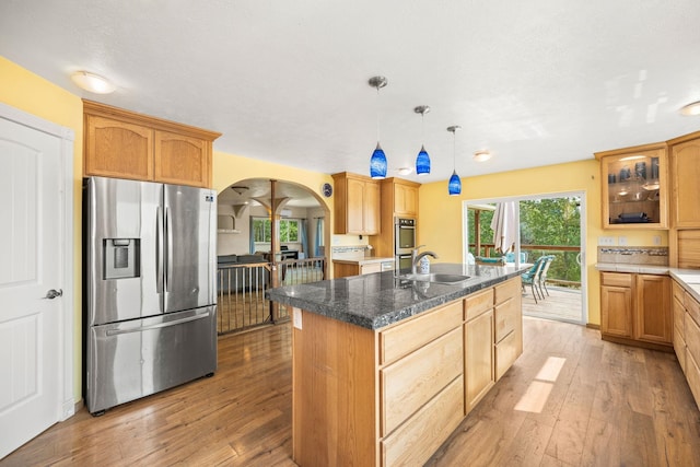 kitchen with a sink, a kitchen island with sink, stainless steel refrigerator with ice dispenser, light wood finished floors, and arched walkways
