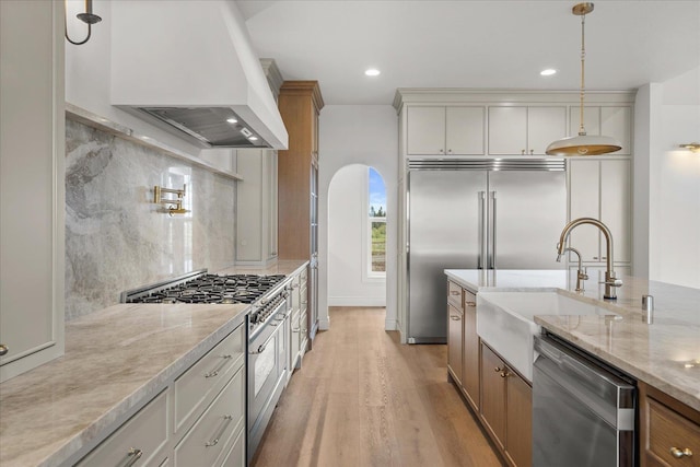 kitchen with high quality appliances, custom exhaust hood, light wood-type flooring, a sink, and hanging light fixtures