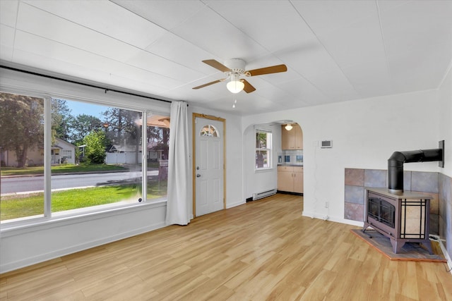 unfurnished living room featuring baseboard heating, baseboards, ceiling fan, a wood stove, and light wood-type flooring