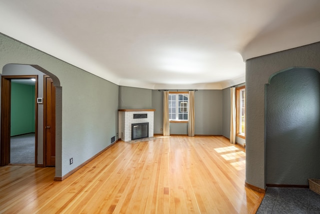 unfurnished living room featuring visible vents, a textured wall, a brick fireplace, arched walkways, and light wood-style floors
