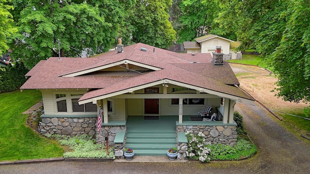 view of front facade featuring a shingled roof, stone siding, covered porch, and a front yard