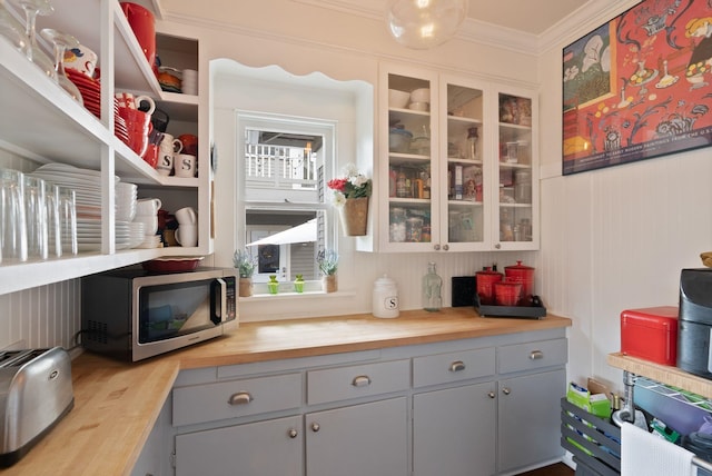 kitchen with stainless steel microwave, butcher block counters, gray cabinetry, crown molding, and glass insert cabinets