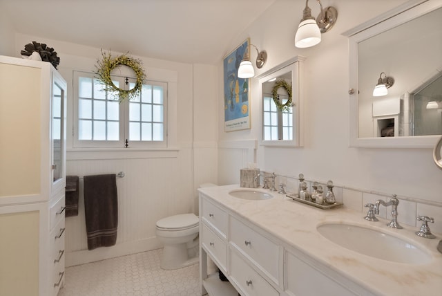 bathroom featuring a wainscoted wall, toilet, double vanity, tile patterned floors, and a decorative wall