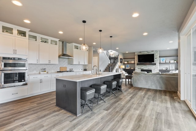 4513 S Crows Nest Ln, Spokane, WA 99206 - kitchen featuring wall chimney range hood, appliances with stainless steel finishes, light wood-style floors, a stone fireplace, and a kitchen bar
