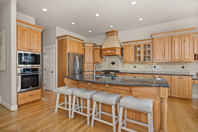 5614 N Thierman Rd, Spokane, WA 99217 - kitchen featuring appliances with stainless steel finishes, custom range hood, backsplash, light wood-style flooring, and a center island with sink