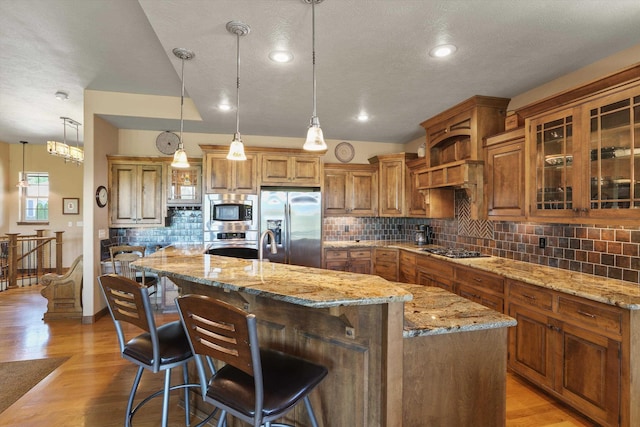 1604 N Rim View St, Spokane, WA 99224 - kitchen with stainless steel appliances, a kitchen island with sink, light stone countertops, brown cabinetry, and decorative backsplash