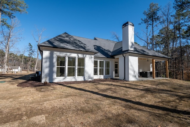 back of house featuring a shingled roof, central AC unit, a chimney, a yard, and brick siding