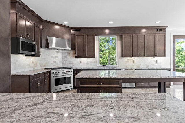 kitchen featuring appliances with stainless steel finishes, light stone countertops, dark brown cabinets, and wall chimney range hood