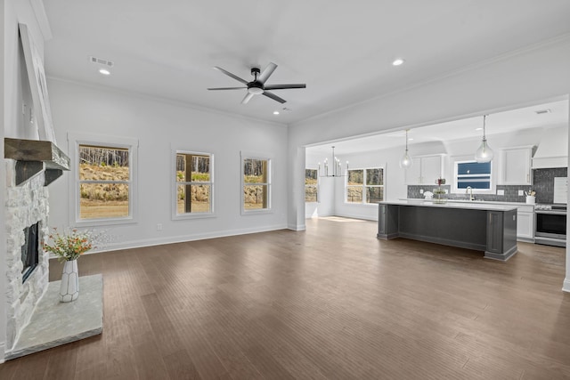 unfurnished living room featuring a sink, crown molding, a stone fireplace, ceiling fan with notable chandelier, and dark wood-style flooring