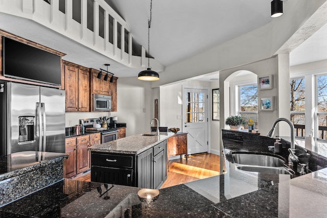 kitchen featuring a kitchen island with sink, stainless steel appliances, a sink, brown cabinets, and dark stone counters