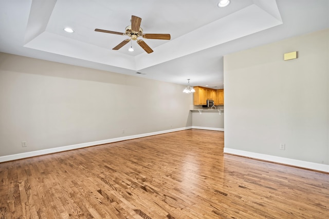 unfurnished living room featuring ceiling fan with notable chandelier, light wood-type flooring, and a tray ceiling