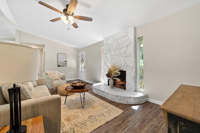 living area with baseboards, lofted ceiling, a ceiling fan, a stone fireplace, and dark wood-type flooring