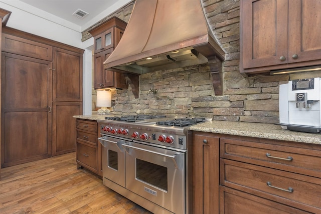 kitchen featuring tasteful backsplash, double oven range, light stone counters, custom range hood, and light hardwood / wood-style flooring