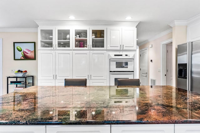 kitchen with crown molding, dark stone countertops, stainless steel fridge, double oven, and white cabinets