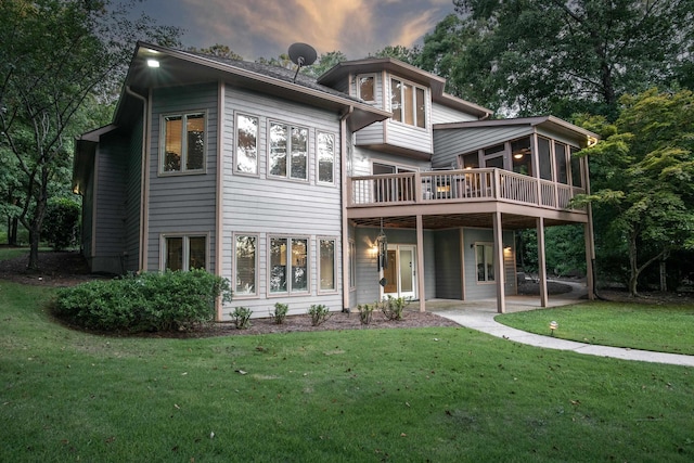 back house at dusk featuring a wooden deck, a patio area, a sunroom, and a lawn