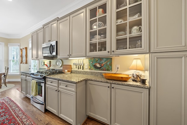 kitchen with dark wood-style flooring, ornamental molding, light stone counters, glass insert cabinets, and appliances with stainless steel finishes