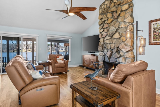 living area featuring a ceiling fan, baseboards, high vaulted ceiling, a stone fireplace, and light wood-style floors