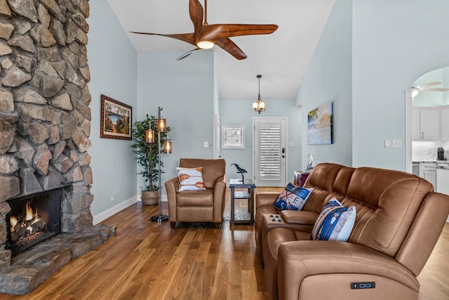 living area featuring wood finished floors, baseboards, high vaulted ceiling, ceiling fan, and a stone fireplace