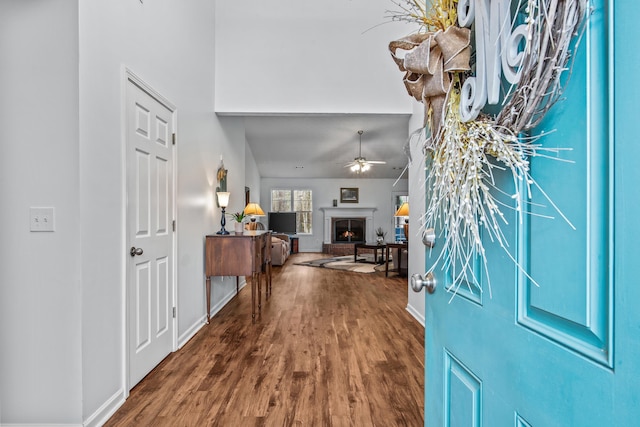 foyer with wood finished floors, a ceiling fan, baseboards, and a warm lit fireplace