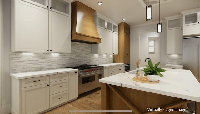 kitchen featuring premium range hood, range with two ovens, hanging light fixtures, a kitchen island with sink, and light stone counters