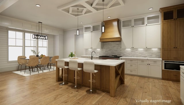 kitchen with an island with sink, white cabinetry, decorative backsplash, custom exhaust hood, and light hardwood / wood-style floors