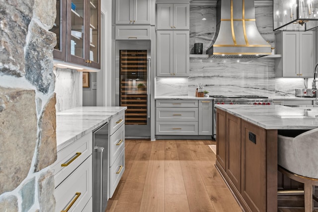 kitchen featuring light wood finished floors, gray cabinetry, light stone countertops, wine cooler, and wall chimney exhaust hood