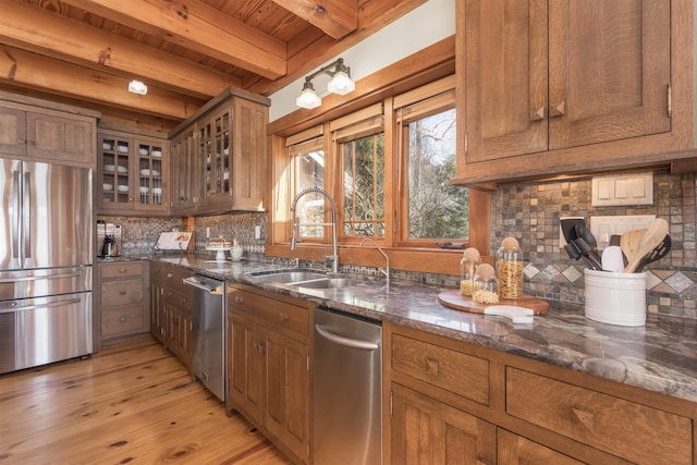 kitchen with brown cabinetry, glass insert cabinets, dark stone countertops, stainless steel appliances, and a sink